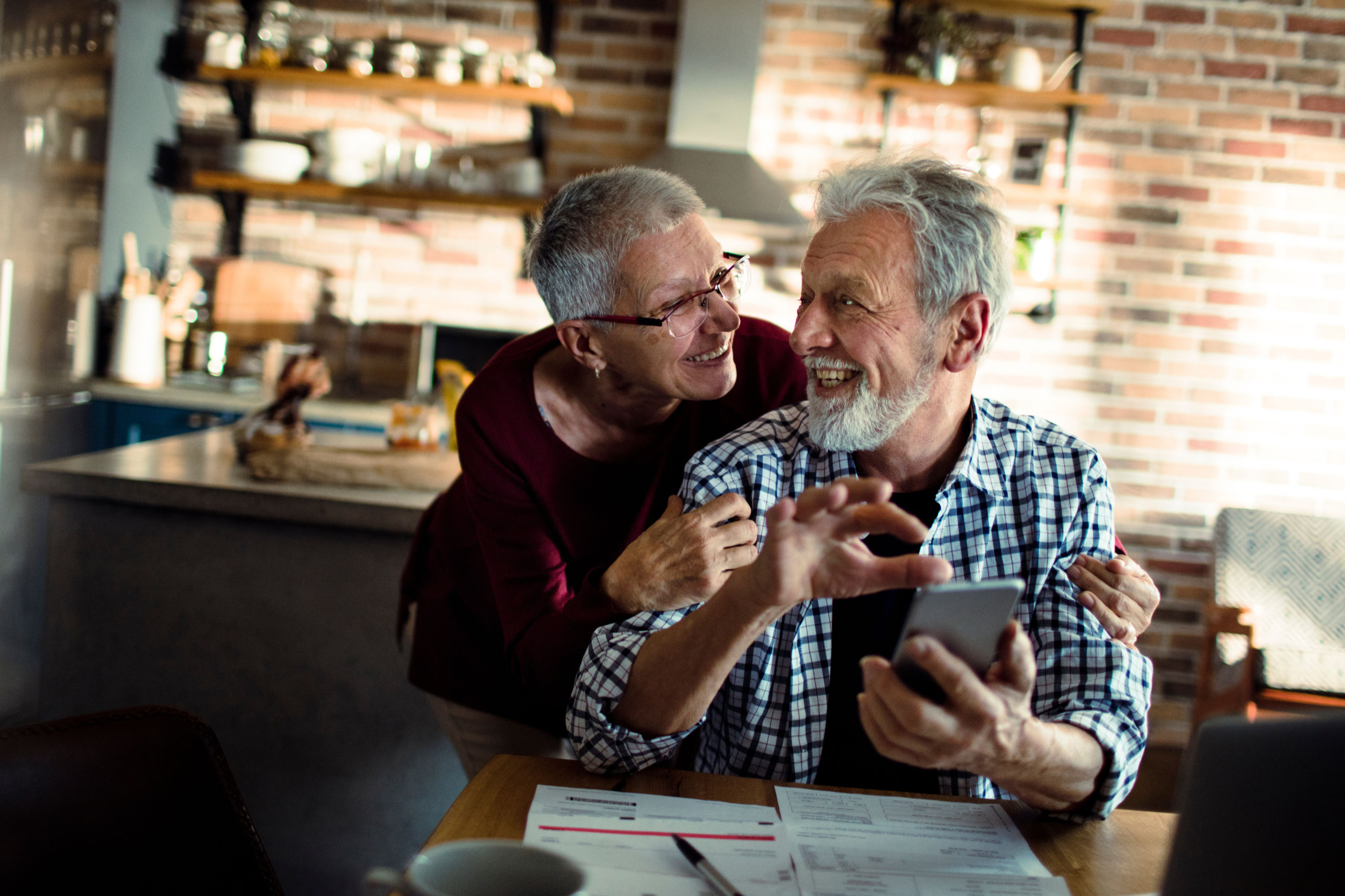 Ein älteres Ehepaar sitzt lachend und glücklich am Küchentisch über Dokumenten und freut sich über die Vorteile der Sozialversicherung.