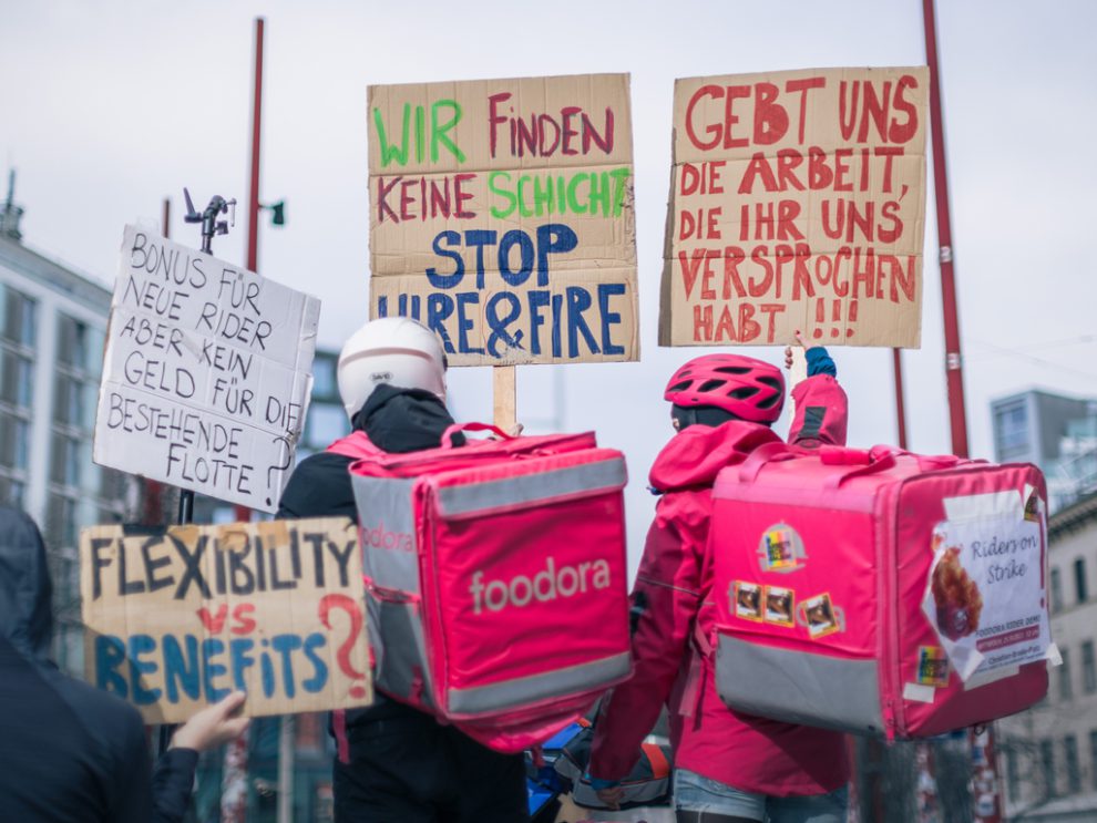 Foodora-Rider:innen demonstrieren auf der Mariahilfer. Auf den Schildern steht "Gebt uns die Arbeit, die ihr versprochen habt", "Wir finden keine Schichten. Stoppt Hire&Fire", "Flexibility vs. Benefits?", "Bonus für neue Rider, aber kein Geld für die bestehende Flotte".