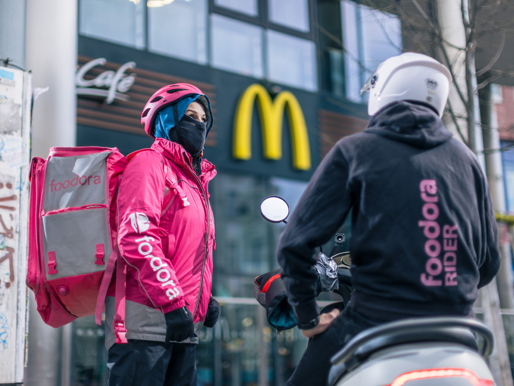 Eine Foodora-Rider:in gekleidet in der Rider:innenuniform steht neben einem Kollegen, der auf einem Motorrad sitzt. Auch er trägt eine Jacke mit dem Logo von Foodora.