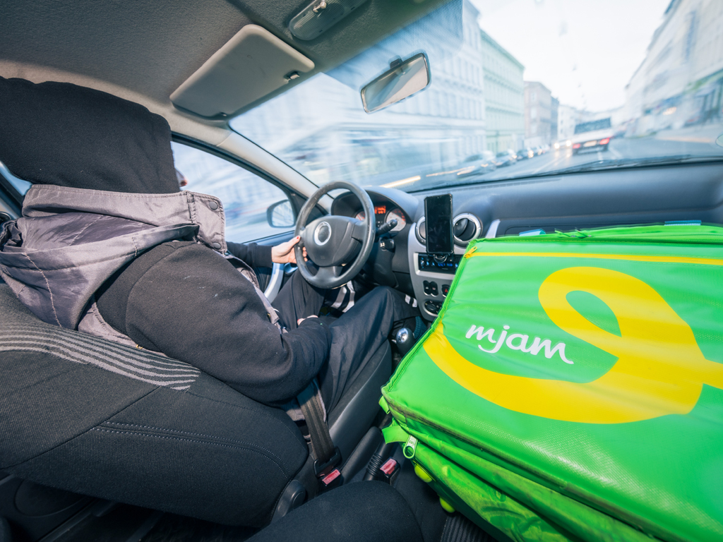 Ein Mann sitzt am Steuer eines Autos und fährt die Straße entlang. Auf dem Beifahrersitzt liegt ein Rucksack mit dem alten Logo von Foodora.