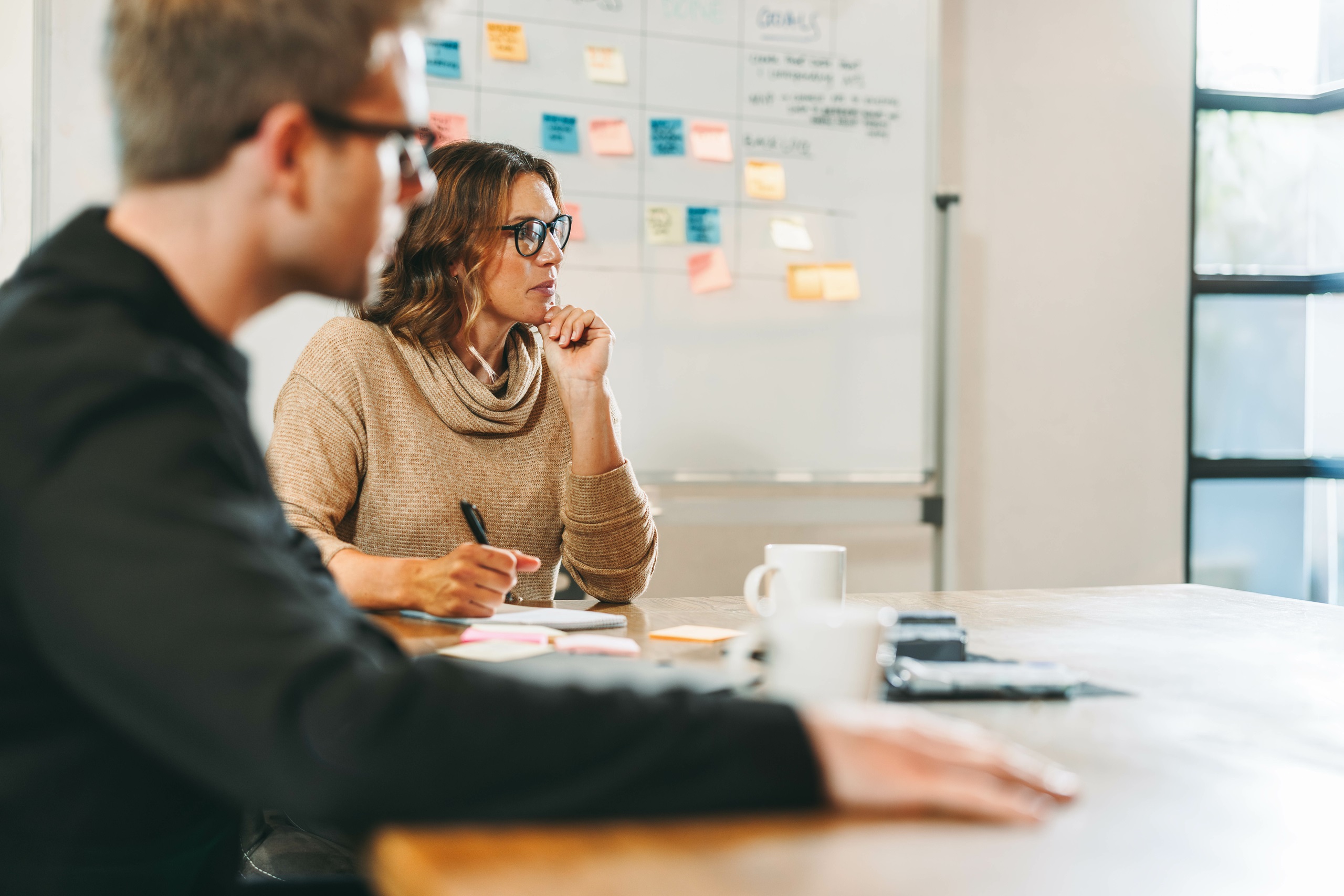 Eine Frau und ein Mann sitzen an einem Tisch, auf dem Tassen stehen. Hinter ihnen ist eine Pinnwand mit bunten Post-Its. Die Frau hält einen Stift in der Hand und hat einen Block vor sich. Sie arbeiten an einem kreativen Projekt. 