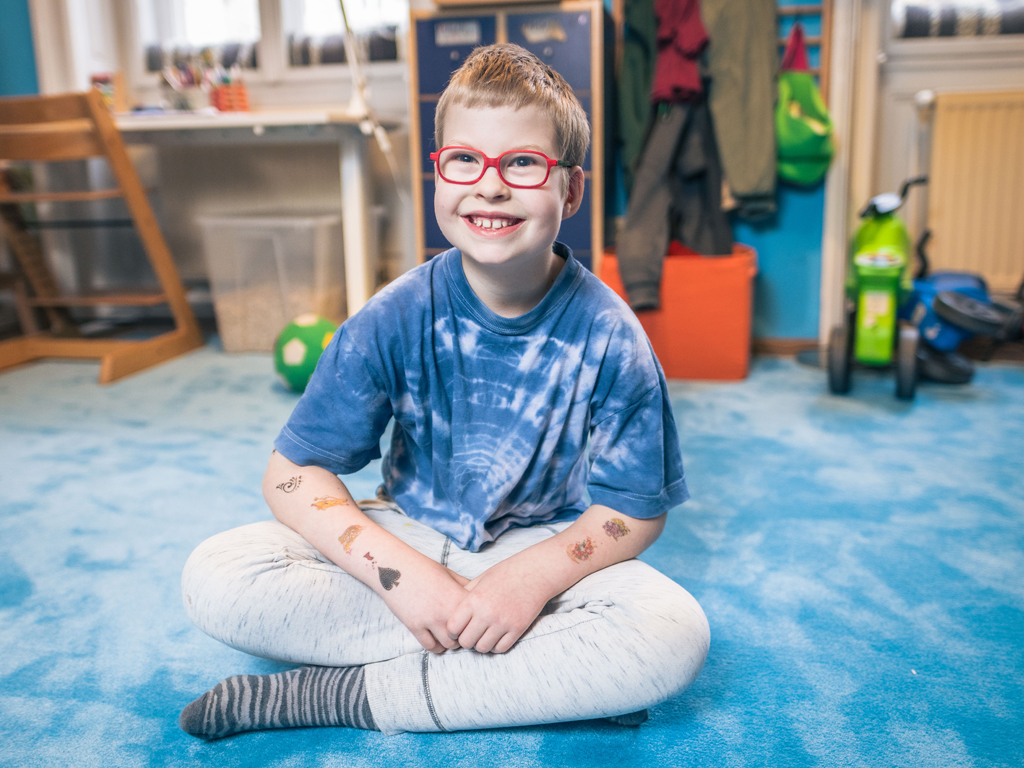 Clemens, ein Kind mit Behinderung, sitzt glücklich auf einem Teppich einer Bildungseinrichtung mit inklusivem Lernen. 
