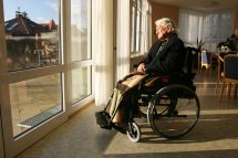 Eine alte Frau sitzt in einem Rollstuhl und schaut aus einem Fenster raus. Symbolbild für das Schmerzmanagement in Pflegeheimen.