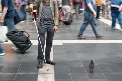 Ein blinder Mann mit Stock in einem belebten Bahnhof mit Barrierefreiheit.