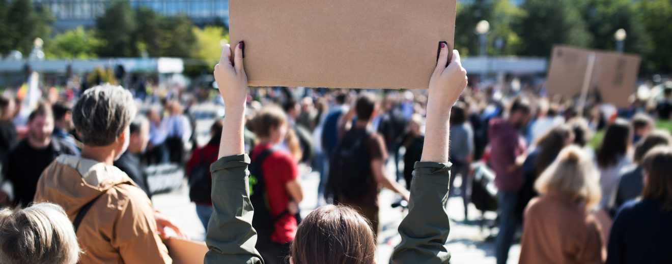 Menschen bei einer Demonstration. Eine Frau hält ein Plakat hoch. Symbolbild für den Kommentar von Kid Pex.