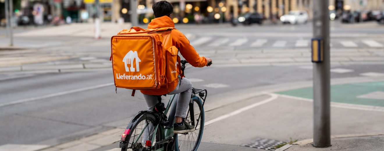 Ein Lieferando-Fahrradbote mit typischer orangefarbener Jacke wartet bis die Ampel grün wird