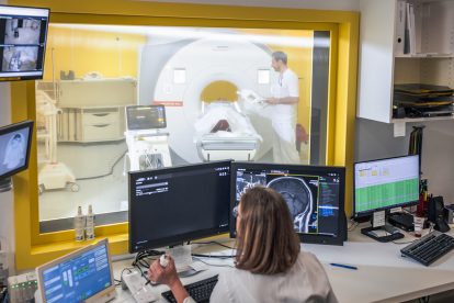 Eine Frau sitzt vor einem PC. Hinter ihr liegt eine Person vor einer MRT-Röhre. Symbolbild für Arbeitssicherheit und Digitalisierung.
