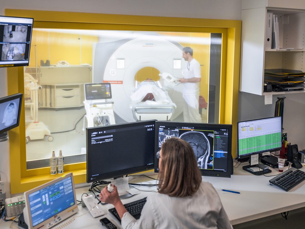 Eine Frau sitzt vor einem PC. Hinter ihr liegt eine Person vor einer MRT-Röhre. Symbolbild für Arbeitssicherheit und Digitalisierung.