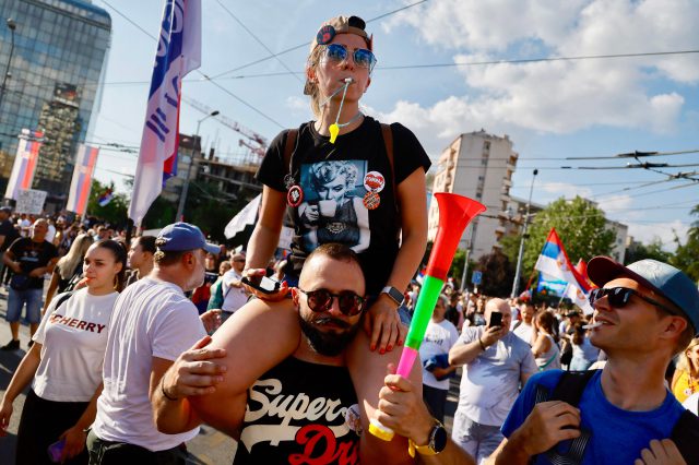 Eine Frau bläst in eine Trillerpfeife und sitzt auf den Schultern eines Mannes bei einem Protest gegen das Vučić-Regime.