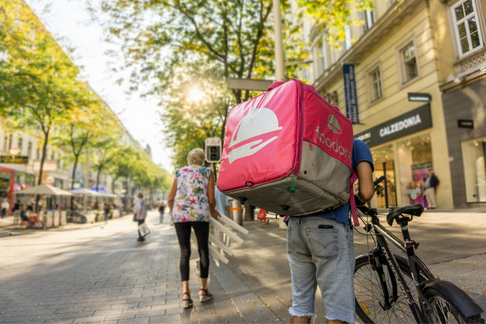 Ein Mann mit einem Foodora-Rucksack steht auf der Mariahilfer neben seinem Fahrrad.