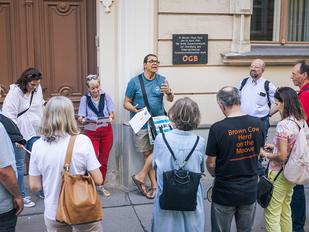 Ein Mann steht vor einem Gebäude. Auf dem Gebäude ist eine Plakette angebracht mit den Worten: "In diesem Haus fand am 13. April 1945 die erste Zusammenkunft des Österreichischen Gewerkschaftsbundes statt. ÖGB". Der Mann spricht zu einer Gruppe von Menschen, die teilweise einen Flyer lesen.