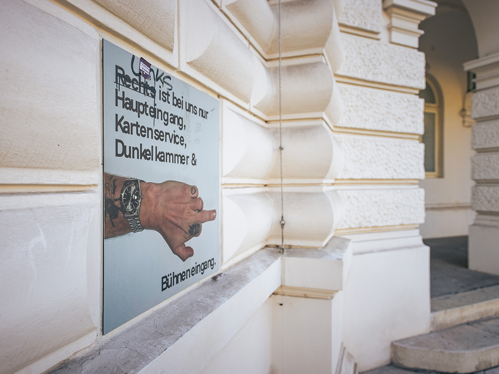 Eine Wand des Wiener Volkstheaters. Darauf steht "Rechts ist bei uns nur Haupteingang, Kartenservice, Dunkelkammer und Bühneneingang". Das Rechts ist durchstrichen, darüber steht Links. Das I von Links ist mit einem Sticker überklebt.