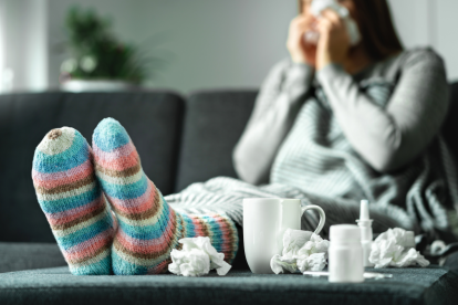 Eine kranke Frau sitzt auf der Couch mit Medikamenten, Tee und Taschentüchern. Symbolbild für den Druck von Arbeitgeber:innen auf Menschen im Krankenstand.