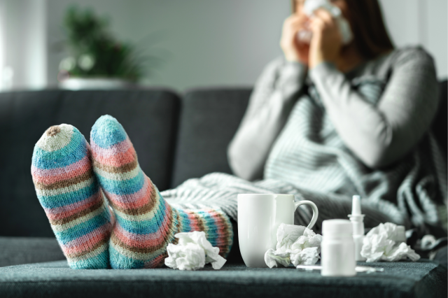 Eine kranke Frau sitzt auf der Couch mit Medikamenten, Tee und Taschentüchern. Symbolbild für den Druck von Arbeitgeber:innen auf Menschen im Krankenstand.