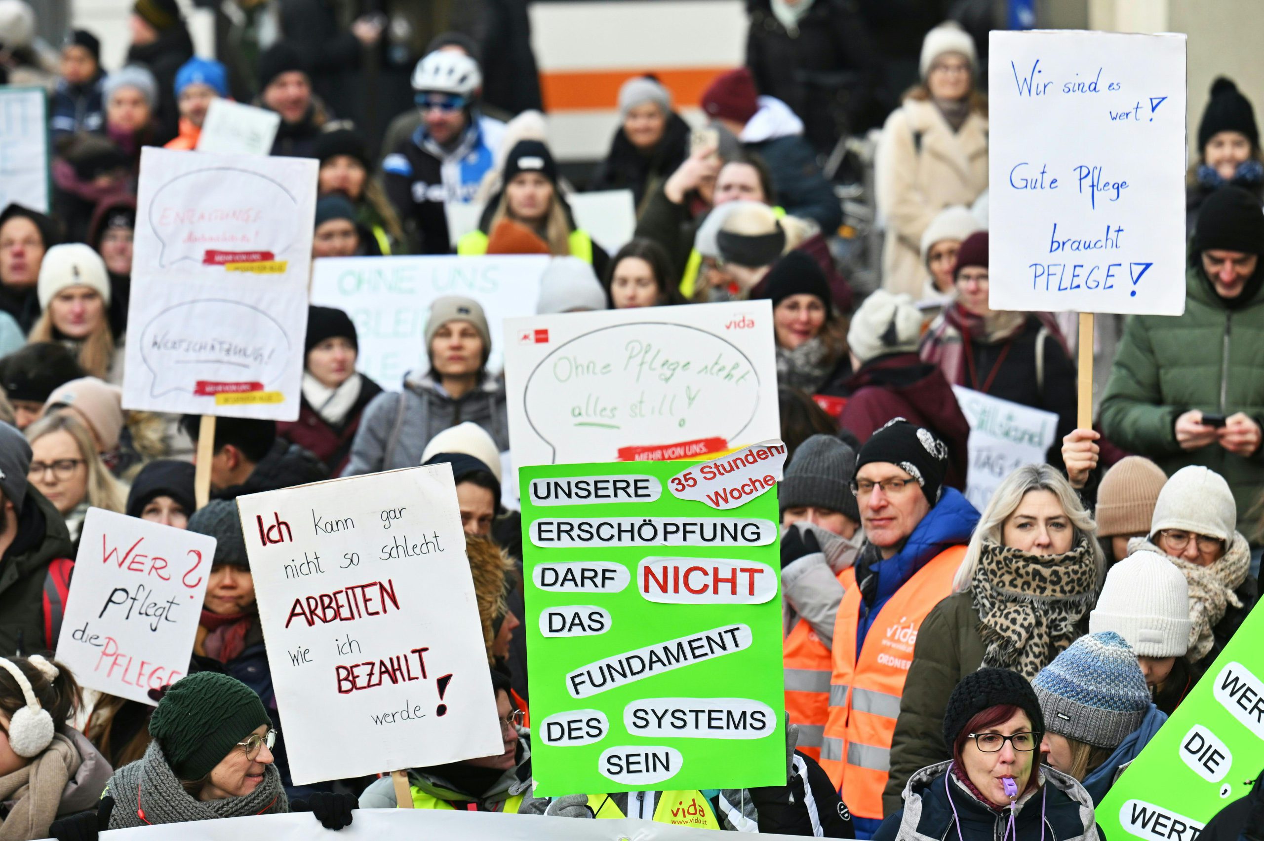 Menschen beim Streik. Sie halten Schilder in die Luft. Darauf steht: "Wer pflegt die Pflege?", "Ich kann gar nicht so schlecht arbeiten, wie ich bezahlt werde", "Unsere Erschöpfung darf nicht das Fundament des Systems sein", "Ohne Pflege steht alles still", "Wir sind es wert! Gute Pflege braucht Pflege!"