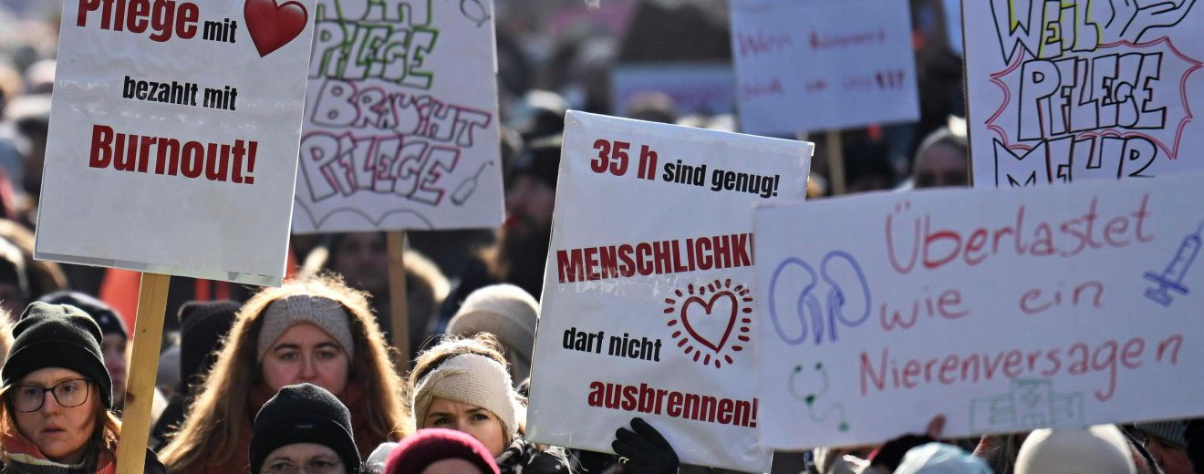 Menschen beim Streik mit Schildern in der Hand. Darauf steht "Pflege mit Herz bezahlt mit Burnout", "Auch Pflege braucht Pflege", 35h sind genug! Menschlichkeit darf nicht ausbrennen", "Überlastet wie ein Nierenversagen"