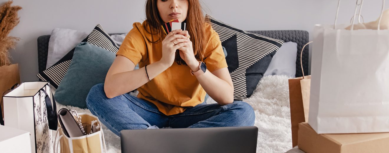 Eine Frau sitzt vor dem Laptop. Neben ihr sind viele Einkaufssackerl. Sie hält eine Kreditkarte in der Hand.