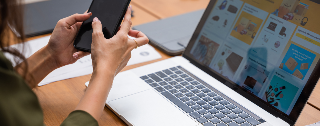 Eine Frau hält ein Handy in der Hand. Vor ihr ist ein Laptop und darauf ist die Website eines Onlinehändlers zu sehen. Symbolbild für Risiken beim Onlineshopping.