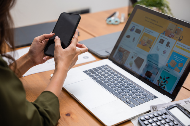 Eine Frau hält ein Handy in der Hand. Vor ihr ist ein Laptop und darauf ist die Website eines Onlinehändlers zu sehen. Symbolbild für Risiken beim Onlineshopping.