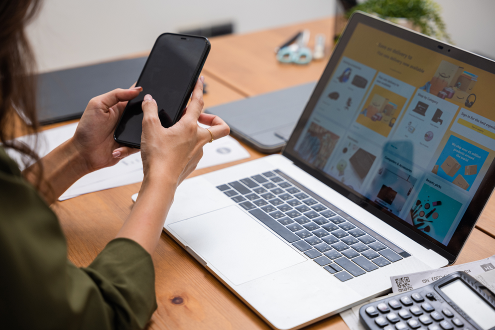 Eine Frau hält ein Handy in der Hand. Vor ihr ist ein Laptop und darauf ist die Website eines Onlinehändlers zu sehen. Symbolbild für Risiken beim Onlineshopping.