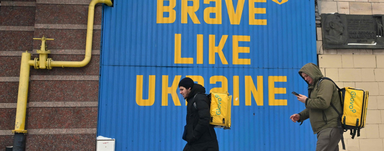 Lieferanten gehen während eines Stromausfalls im Zentrum von Kiew an einem laufenden Stromgenerator unter einem riesigen ukrainischen Kriegs-Slogan vorbei. Symbolbild: Die Rechte von Arbeitnehmer:innen in der Ukraine stehen zunehmend unter Druck. Die Gewerkschaften stehen vor großen Herausforderungen.