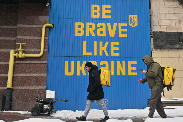 Lieferanten gehen während eines Stromausfalls im Zentrum von Kiew an einem laufenden Stromgenerator unter einem riesigen ukrainischen Kriegs-Slogan vorbei. Symbolbild: Die Rechte von Arbeitnehmer:innen in der Ukraine stehen zunehmend unter Druck. Die Gewerkschaften stehen vor großen Herausforderungen.