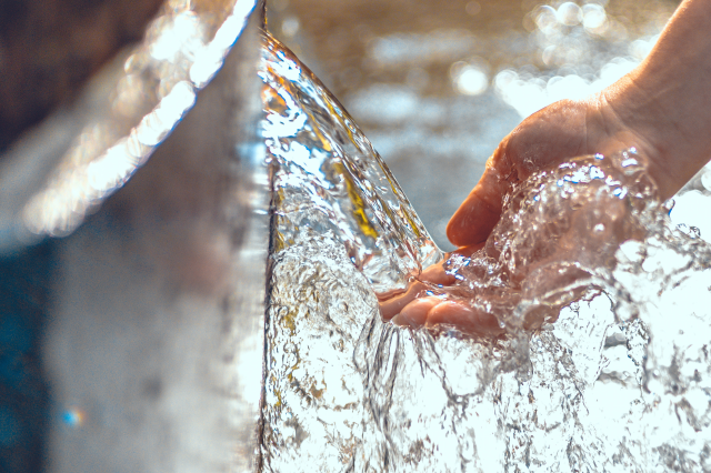Eine Hand berührt einen Wasserstrahl. Symbolbild: Das Wasser könnte knapp werden. Aber wer verbraucht am meisten Wasser? Und wie verteilt man es im Ernstfall?