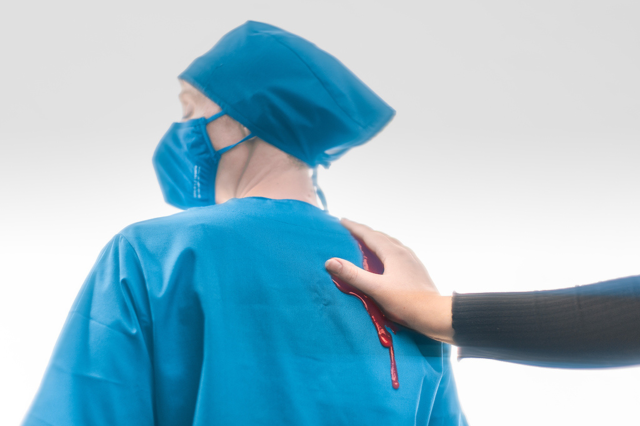 Eine Hand schmiert Blut auf die Uniform einer Pflegeperson. Symbolbild: ÖGB-Expert:innen Claudia Neumayer-Stickler und Julia Stroj möchten die Debatte um die Kassenfusion wieder ankurbeln.
