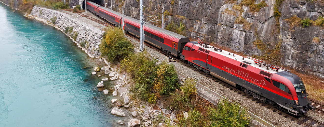 Ein roter Zug fährt aus einem Tunnel neben einem Fluss. Symbolbild für die Mobilitätswende und die Autoindustrie, die immer mehr unter Druck gerät.