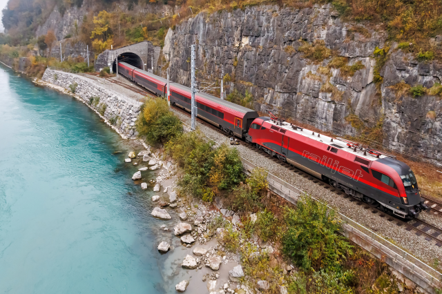 Ein roter Zug fährt aus einem Tunnel neben einem Fluss. Symbolbild für die Mobilitätswende und die Autoindustrie, die immer mehr unter Druck gerät.