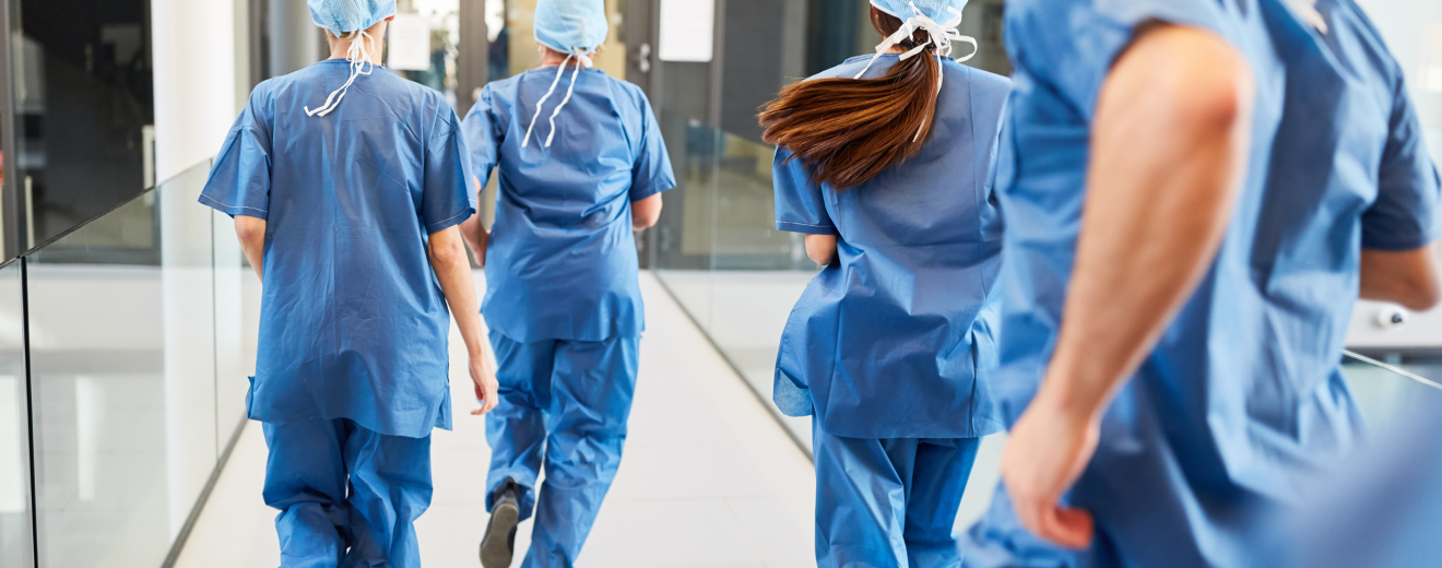Ärzt:innen laufen durch ein Krankenhaus. Sie tragen blaue Scrubs. Symbolbild: Das Gesundheitssystem braucht dringend Reformen um bessere Arbeitsbedingungen und keine Zwei-Klassen-Medizin zu bieten.
