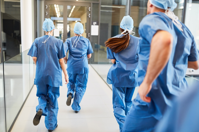 Ärzt:innen laufen durch ein Krankenhaus. Sie tragen blaue Scrubs. Symbolbild: Das Gesundheitssystem braucht dringend Reformen um bessere Arbeitsbedingungen und keine Zwei-Klassen-Medizin zu bieten.