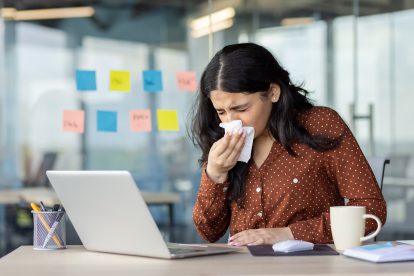 Eine Frau, die an ihrem Schreibtisch im Büro in ein Taschentuch niest, umgeben von Arbeitsunterlagen und einem Laptop. Symbolbild für Krankenstand in Österreich