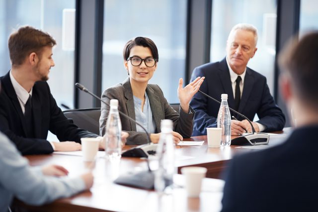 Porträt einer jungen Geschäftsfrau, die während einer Gruppendiskussion in einem Konferenzraum in ein Mikrofon spricht.