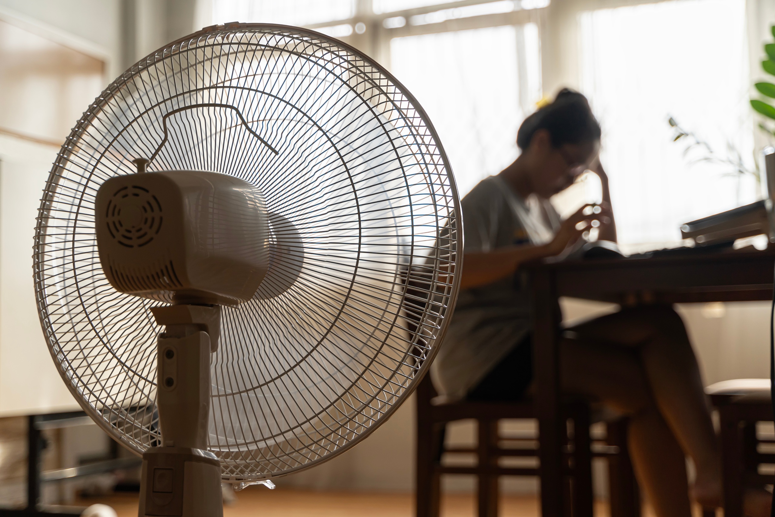 Frau sitzt an einem heißen Sommertag vor einem laufenden Ventilator und leidet unter der Hitze.